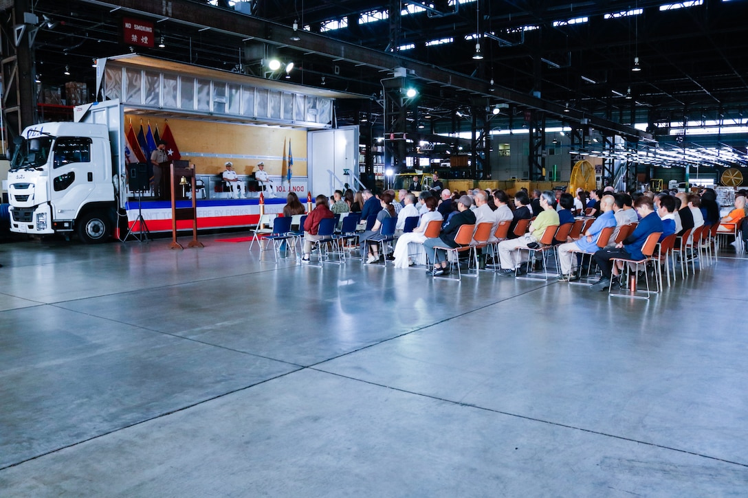 A crowd of people sit in chairs inside a warehouse looking at a stage.