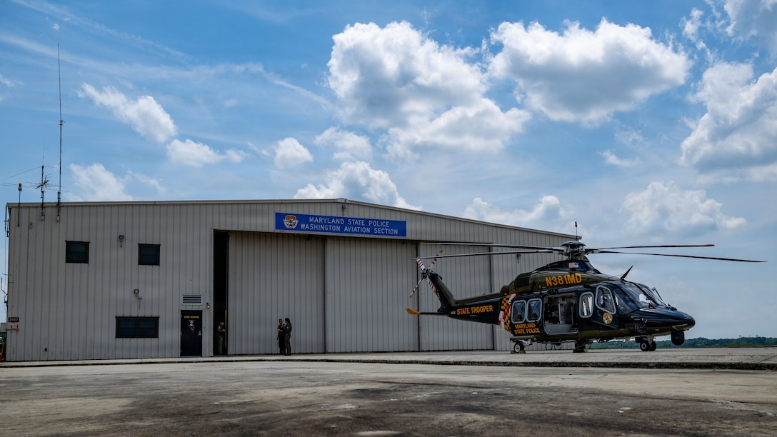 A helicopter sits on a flight pad. There's a hangar in the background.