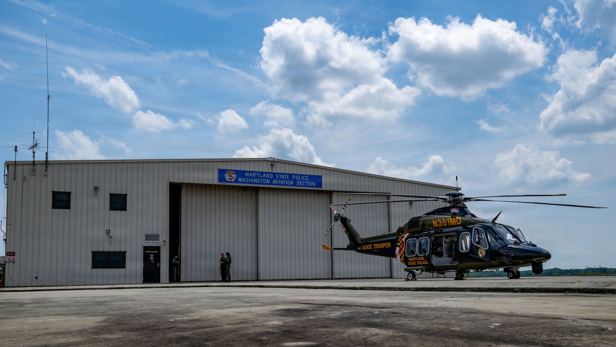 A helicopter sits on a flight pad. There's a hangar in the background.