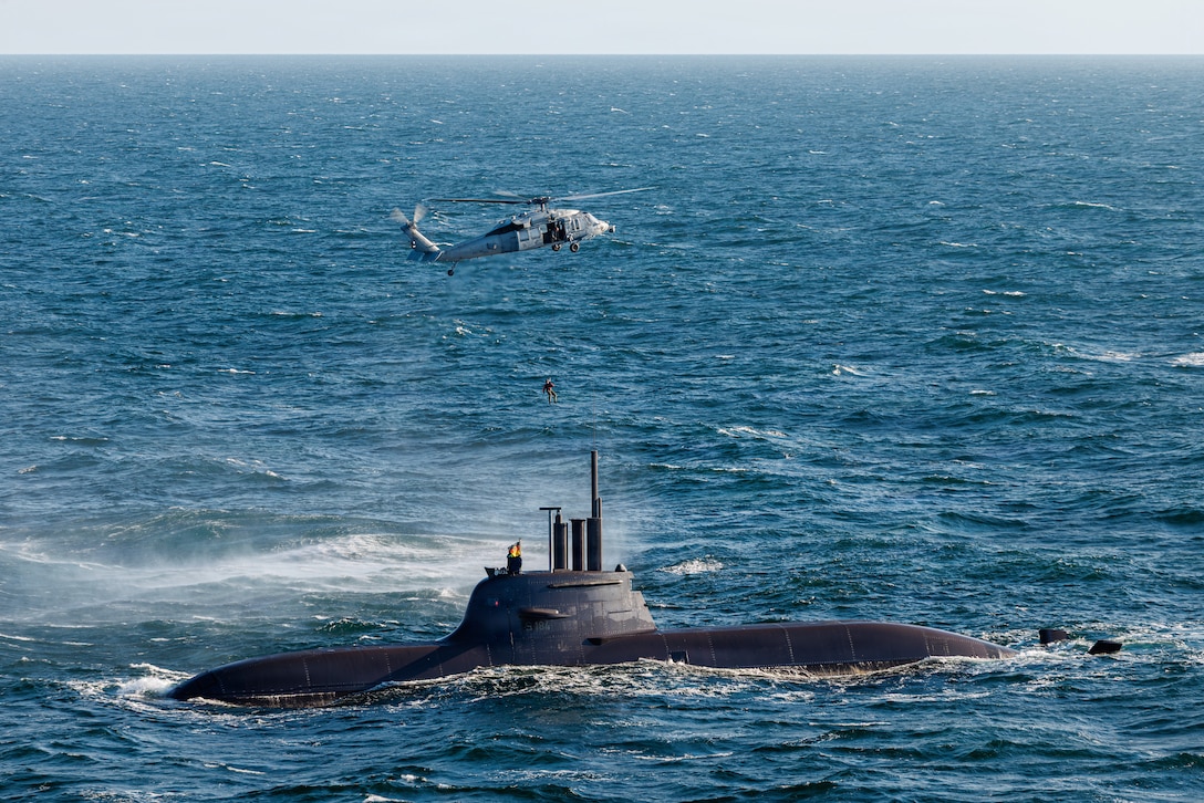 A sailor dangles from a helicopter above a submarine emerging from a choppy sea.