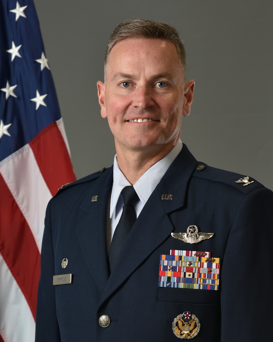 Col Simmons poses for an official photo with a U.S. flag behind him with a grey background.
