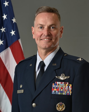 Col Simmons poses for an official photo with a U.S. flag behind him with a grey background.