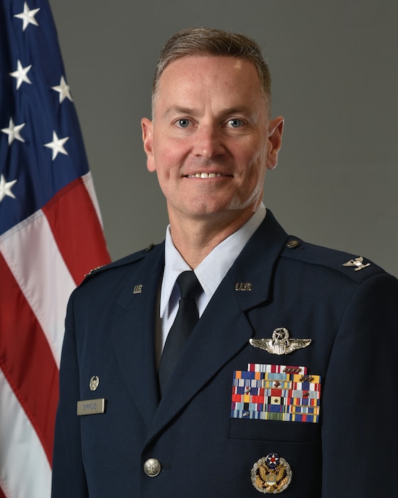 Col Simmons poses for an official photo with a U.S. flag behind him with a grey background.
