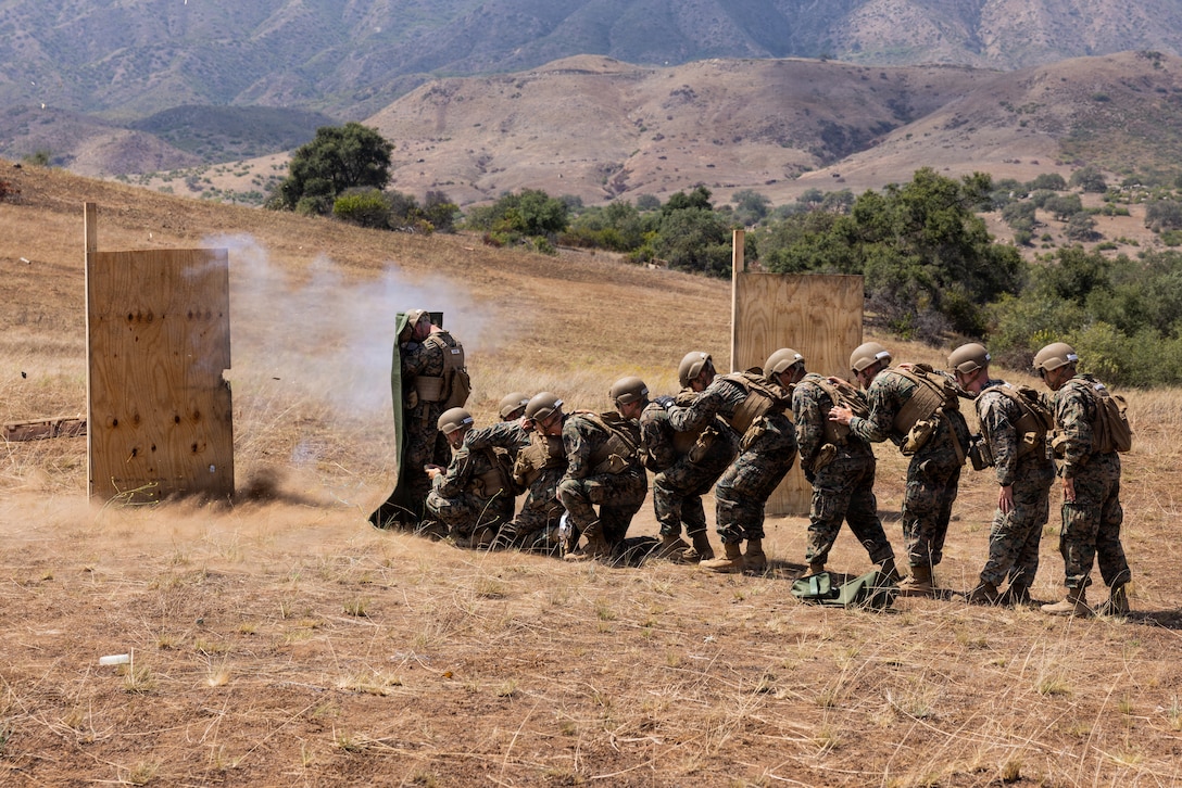 Nine midshipmen wearing camouflage uniforms, protective vests and helmets, are lined up to take cover behind another midshipman in similar gear holding a shield.