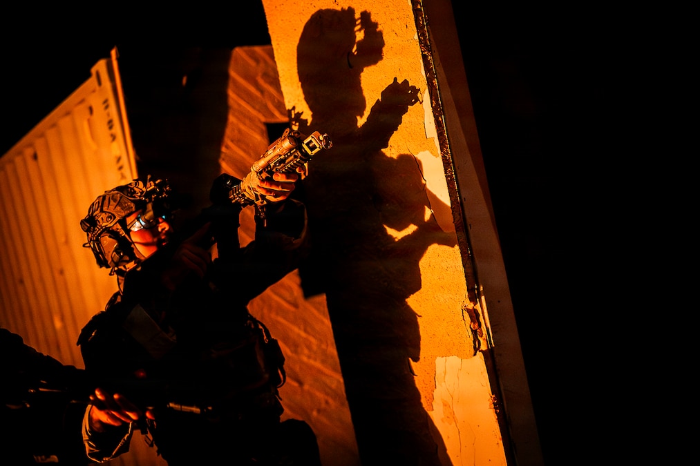 A sailor wearing a helmet and night vision equipment points his rifle around the corner of a structure in a low-light environment.