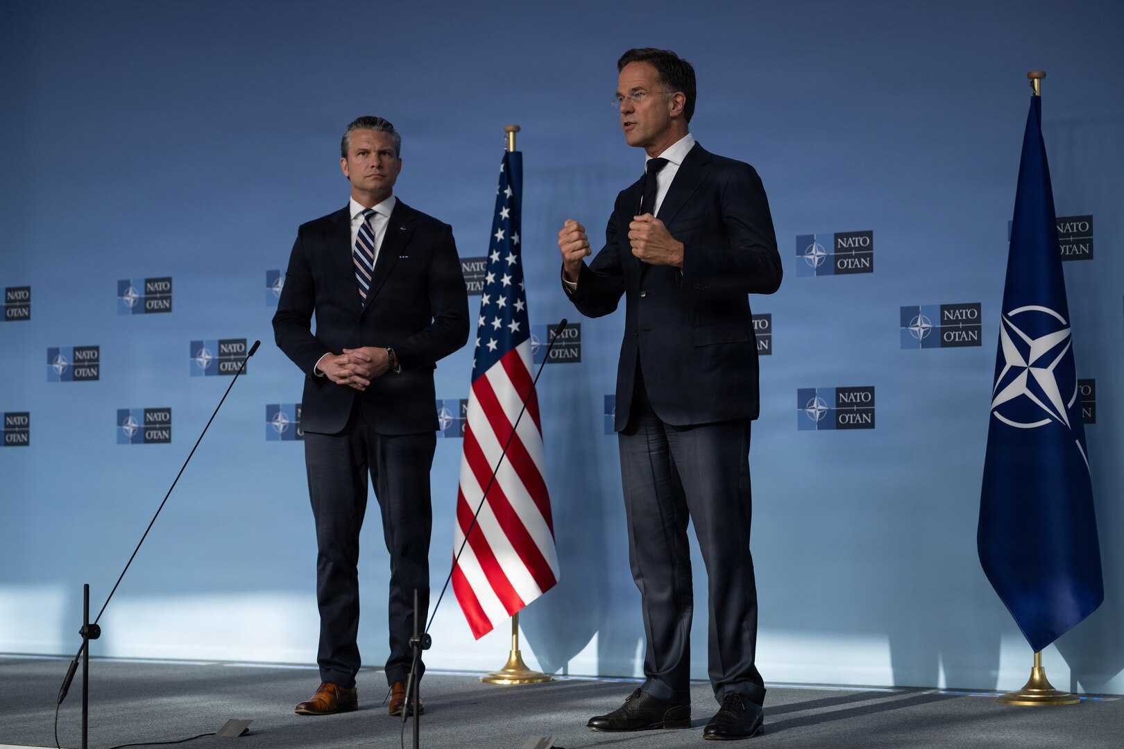 Secretary of War Pete Hegseth stands with another official, who gestures while speaking in front of a blue wall with NATO logos.
