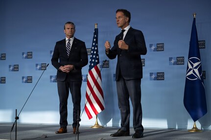 Secretary of War Pete Hegseth stands with another official, who gestures while speaking in front of a blue wall with NATO logos.