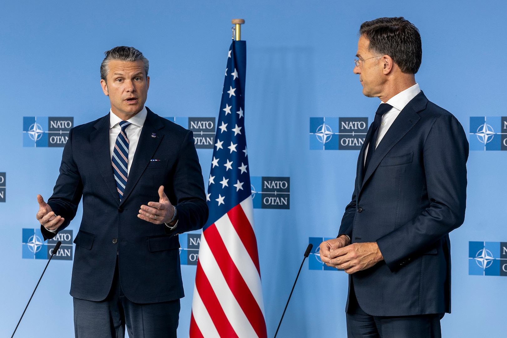 Secretary of War Pete Hegseth speaks while standing with another civilian by a U.S. flag in front of a light blue wall.