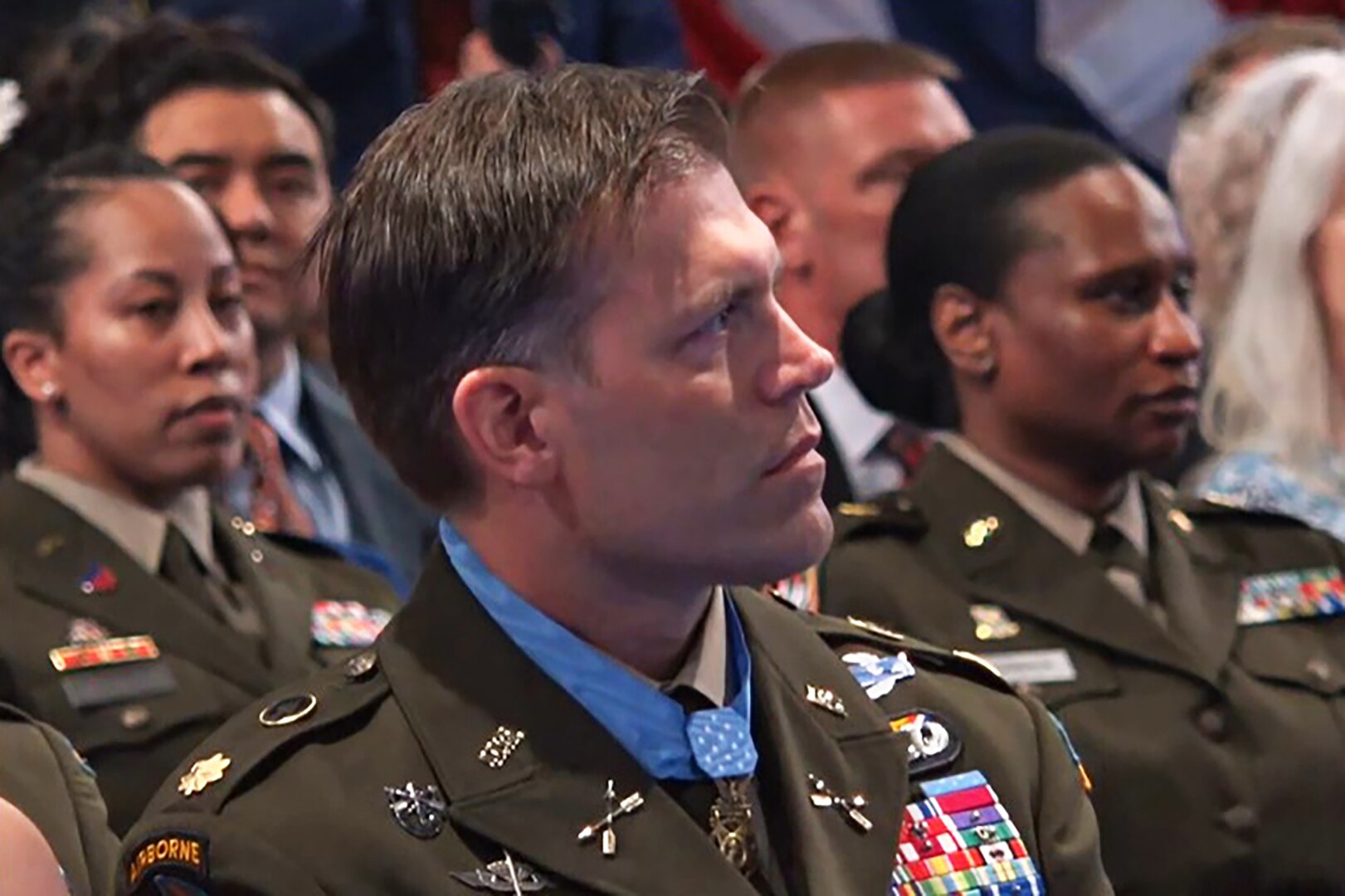 A soldier in a dress uniform speaks at a podium, with service flags behind him.