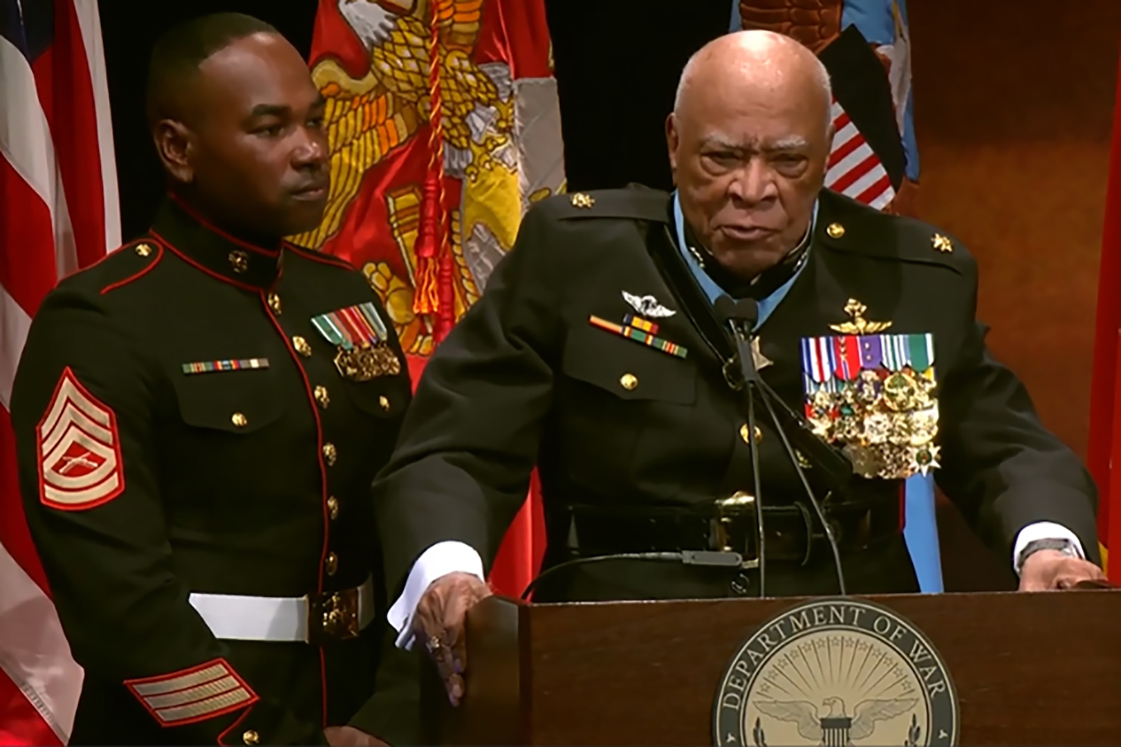A man in a decorated military uniform stands behind a podium , with a Marine standing next to him and service flags in the background.