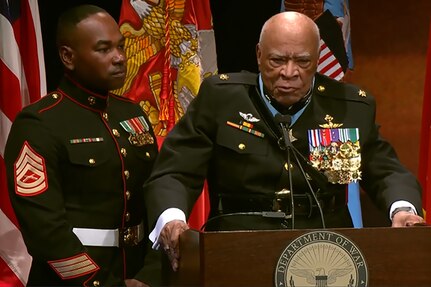 A man in a decorated military uniform stands behind a podium , with a Marine standing next to him and service flags in the background.