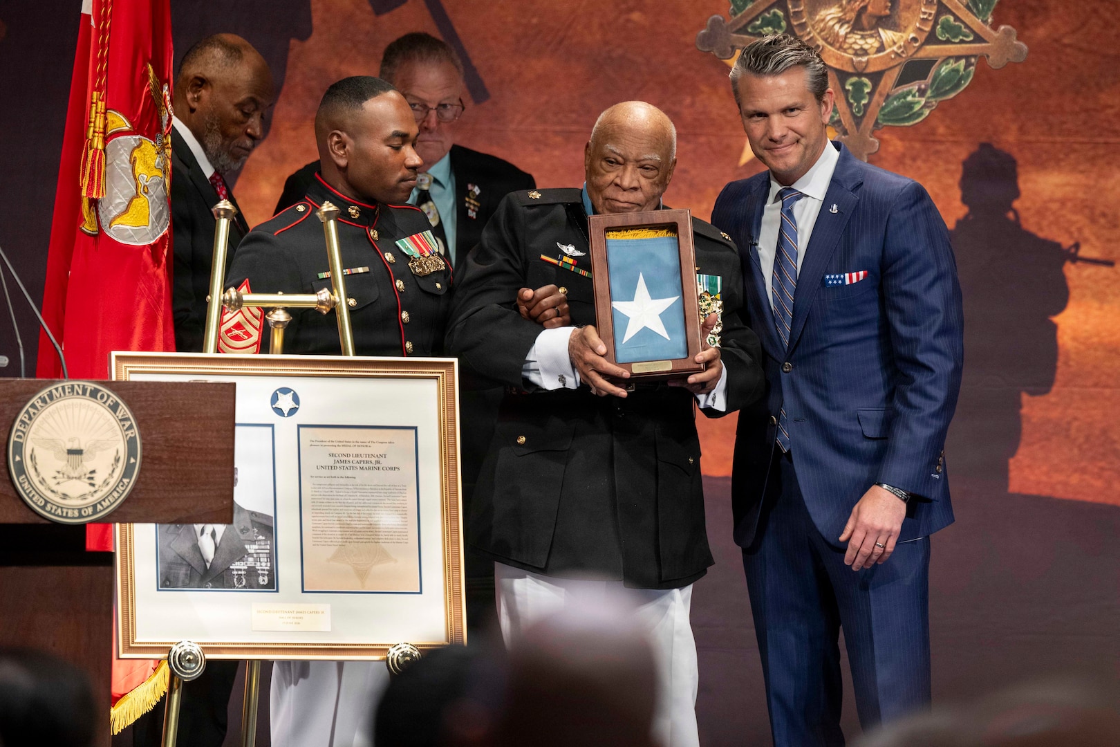 An older man in a military dress uniform and another man in a suit pose for a photo while standing on stage holding up a container with a flag that showcases a five-pointed star; there is another man in a military dress uniform to the side helping the older man stand.