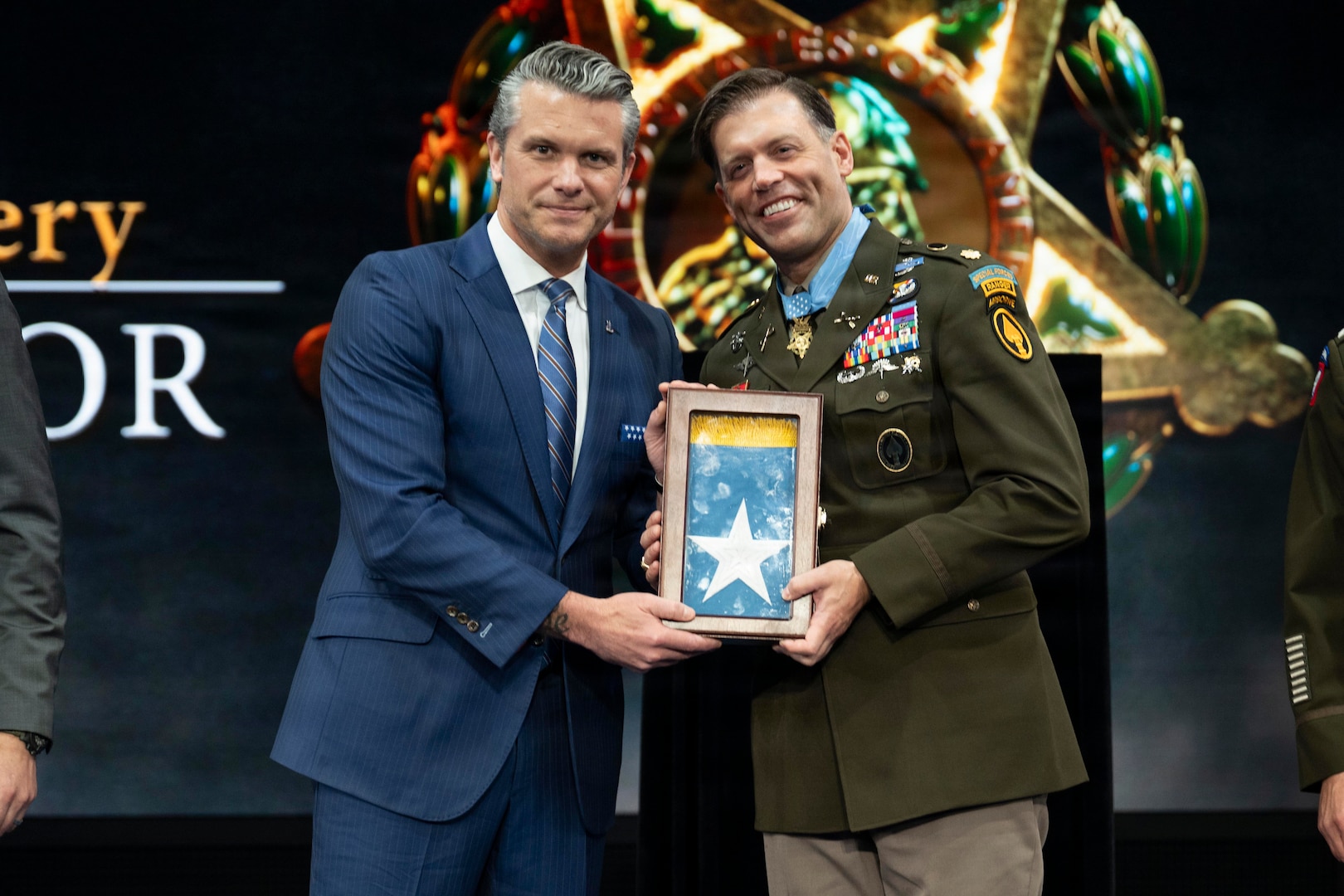 A man in a military dress uniform and another man in a suit pose for a photo on stage while holding up a container with a flag that showcases a five-pointed star.