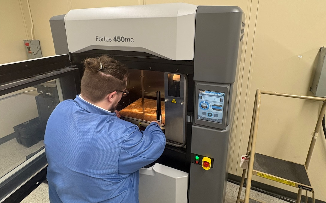 A man with a ponytail, dressed in a blue lab coat reaches into a 3D printing chamber to pull out a 3D printed object. It is a black rod.