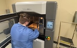 A man with a ponytail, dressed in a blue lab coat reaches into a 3D printing chamber to pull out a 3D printed object. It is a black rod.