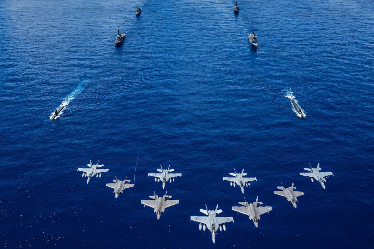 Nine military fighter jets fly in formation while two submarines and four large military ships sail in two rows in the ocean.