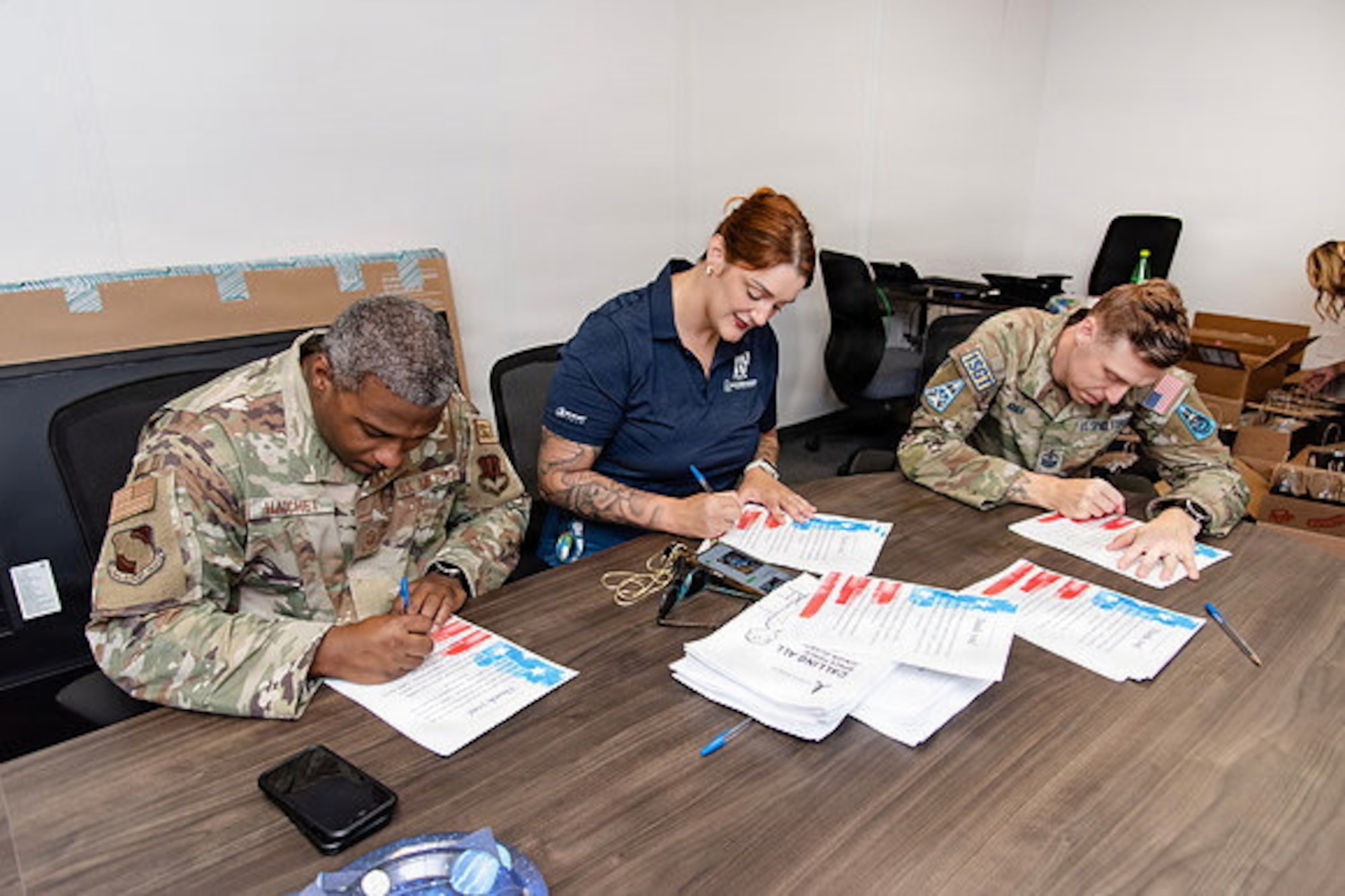 Guardians and volunteers pack boxes of gifts in a room for family members of deployed Guardians. Everyone is either in uniform or civilian clothes and they're in an office/conference space