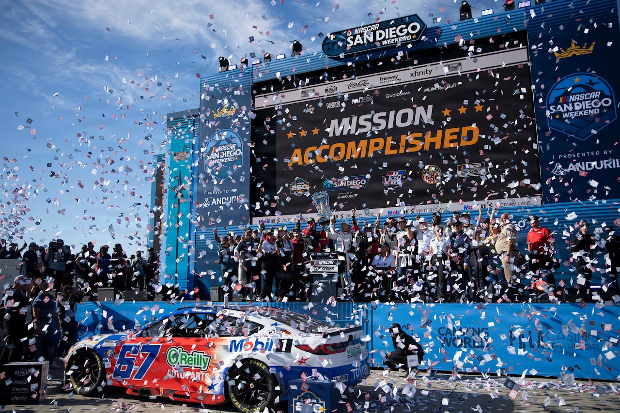 A group of people cheer on a stage as confetti falls onto them; two of the people are holding a large silver trophy. A race car is in front of the stage.