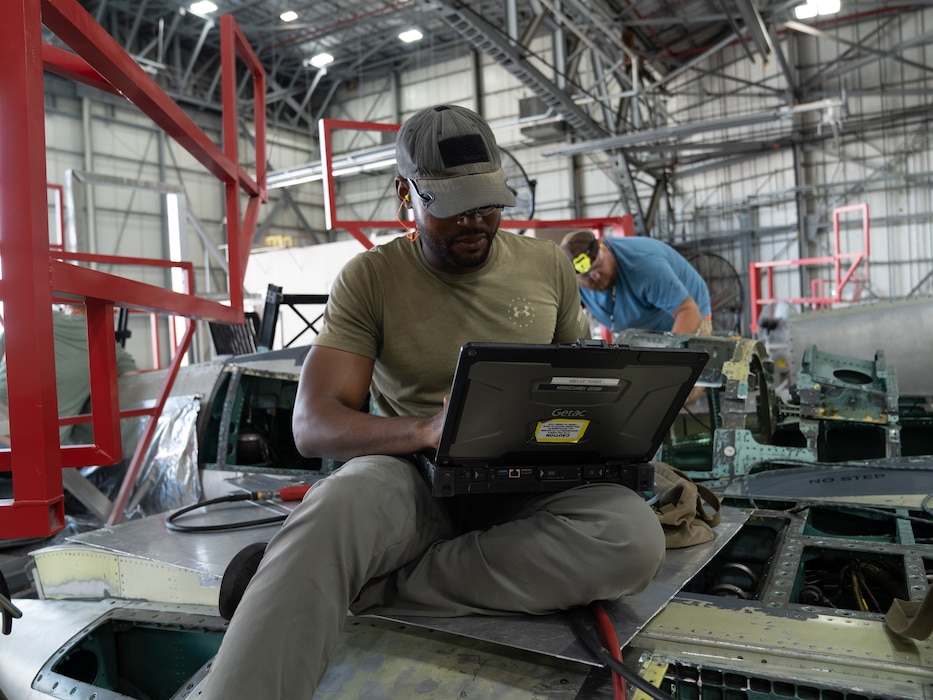 Photo shows man working on laptop sitting atop aircraft frame.