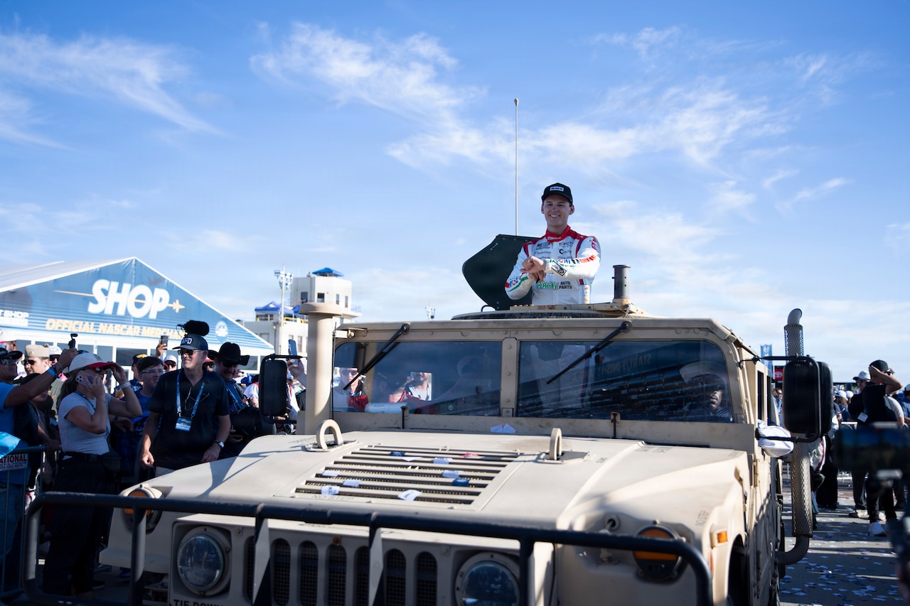 A man in a racing uniform stands in the back of a military vehicle. There is a crowd of people around the vehicle.
