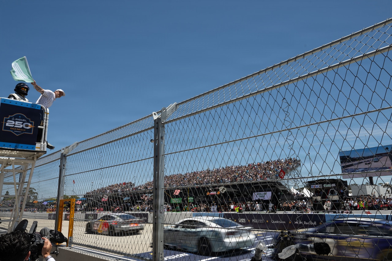A man in a white hat and shirt waves a green flag while standing in an elevated platform above a tall fence, as race cars accelerate on a track below outdoors during daytime; bleachers with spectators are in the background.