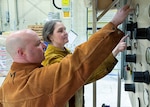 a man in the foreground and a woman next to him both have a single arm up while looking a gauges and pushing buttons on the face of a machine taller than them.