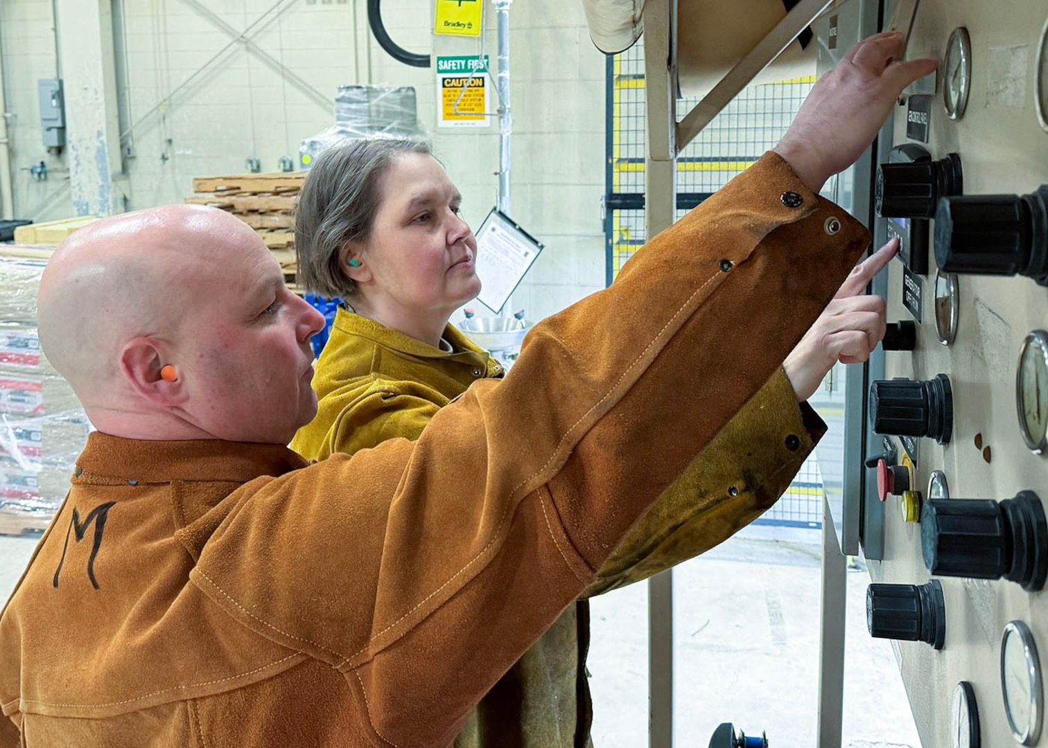a man in the foreground and a woman next to him both have a single arm up while looking a gauges and pushing buttons on the face of a machine taller than them.