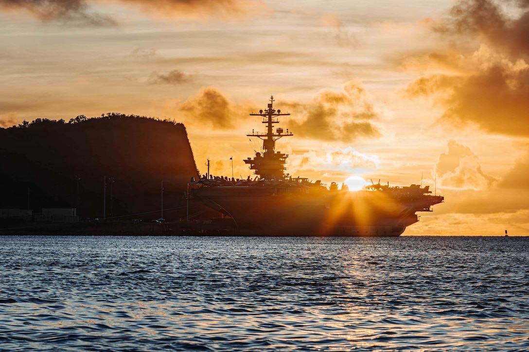 A ship stationed near land is in silhouette, with the sunrise seen above the deck and calm waters in front of it.