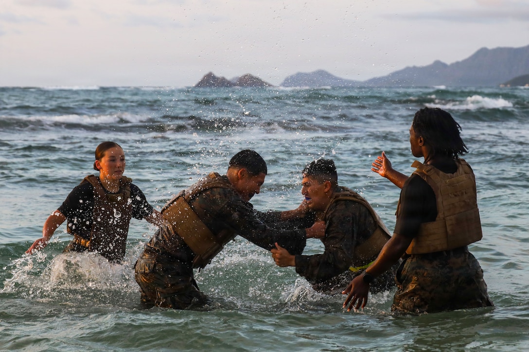 Two Marines grapple in the ocean, with two other Marines standing on either side of them.