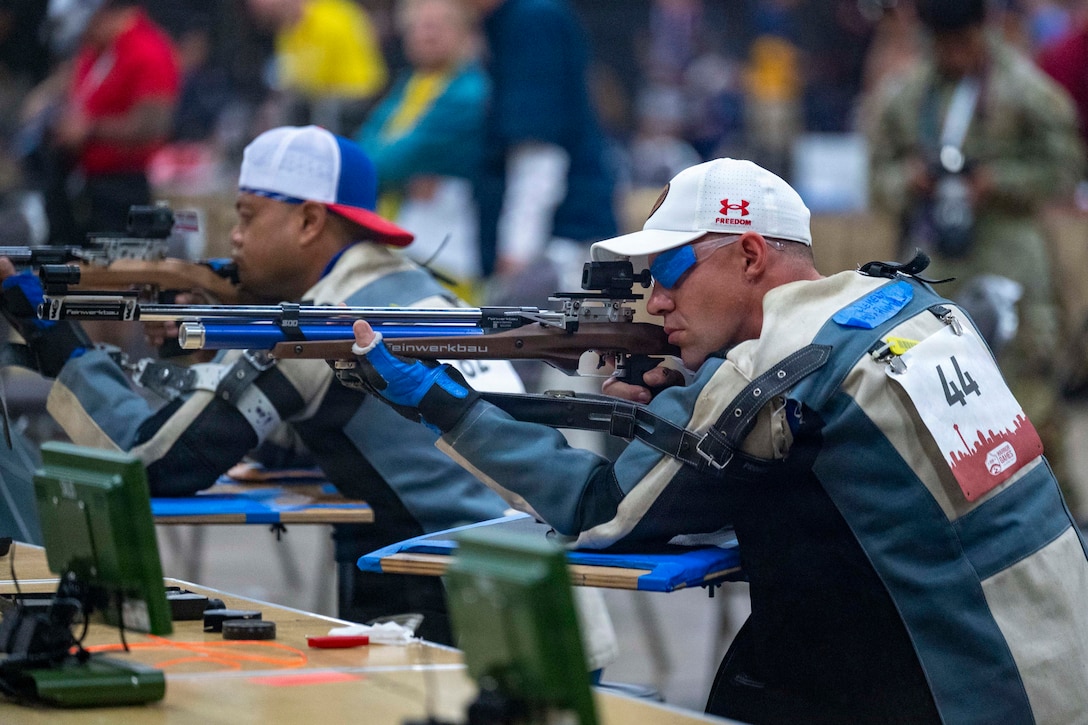 Two people wearing athletic attire and number tags hold rifles steady and aim them, as other people watch in the background.