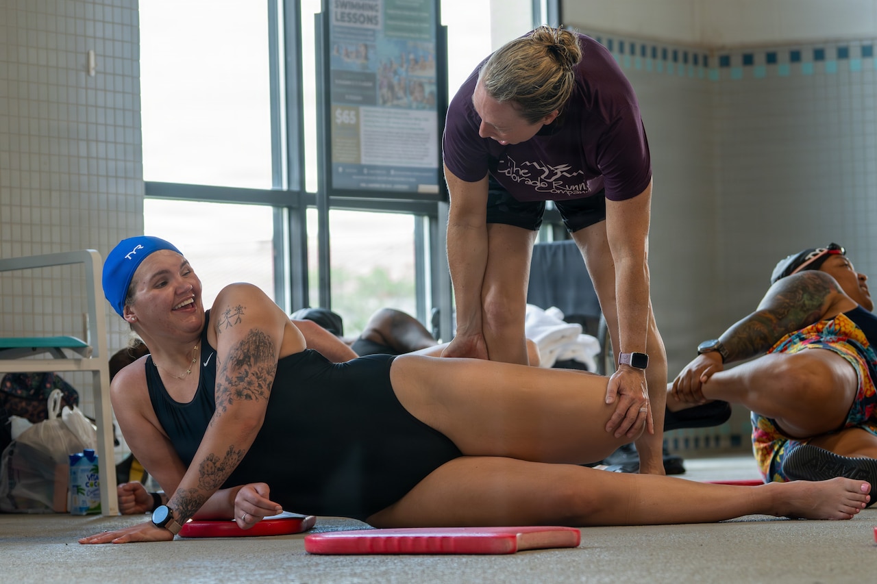 A woman wearing a black bathing suit and swim cap lies on a floor as she is being assisted by another woman in casual attire in an exercise that is lifting her left leg. There is a person also exercising to the right of the women.