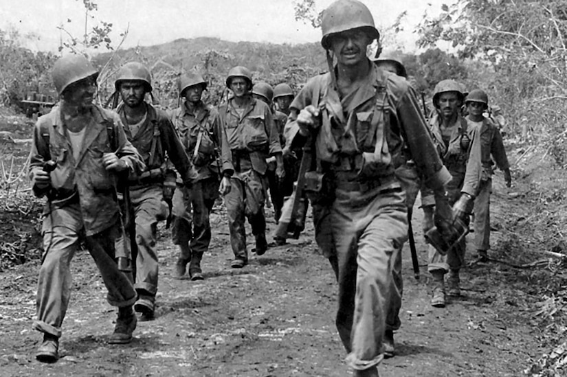 Soldiers walk on a dirt path in a black-and-white photo.