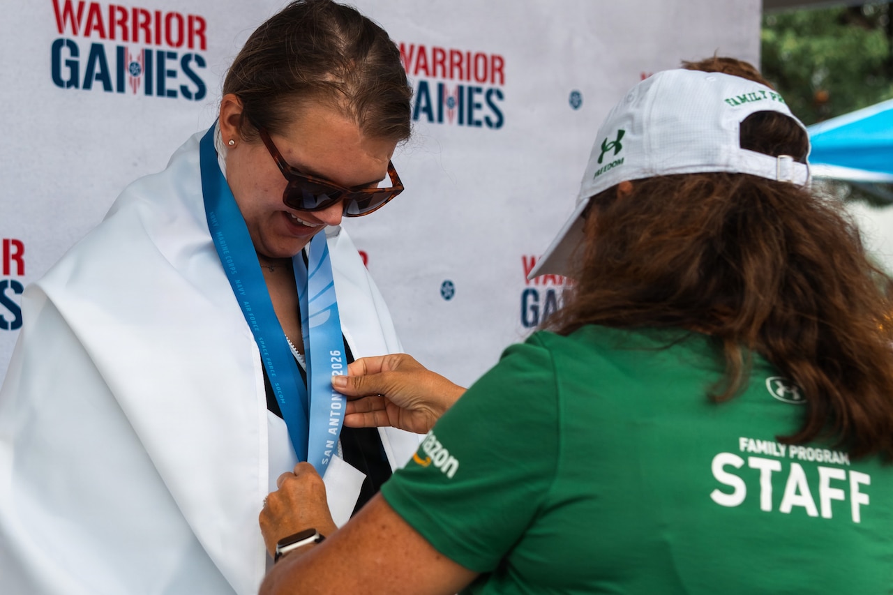 A woman wrapped in a white flag and wearing sunglasses receives a medal on a ribbon from another woman wearing casual attire.