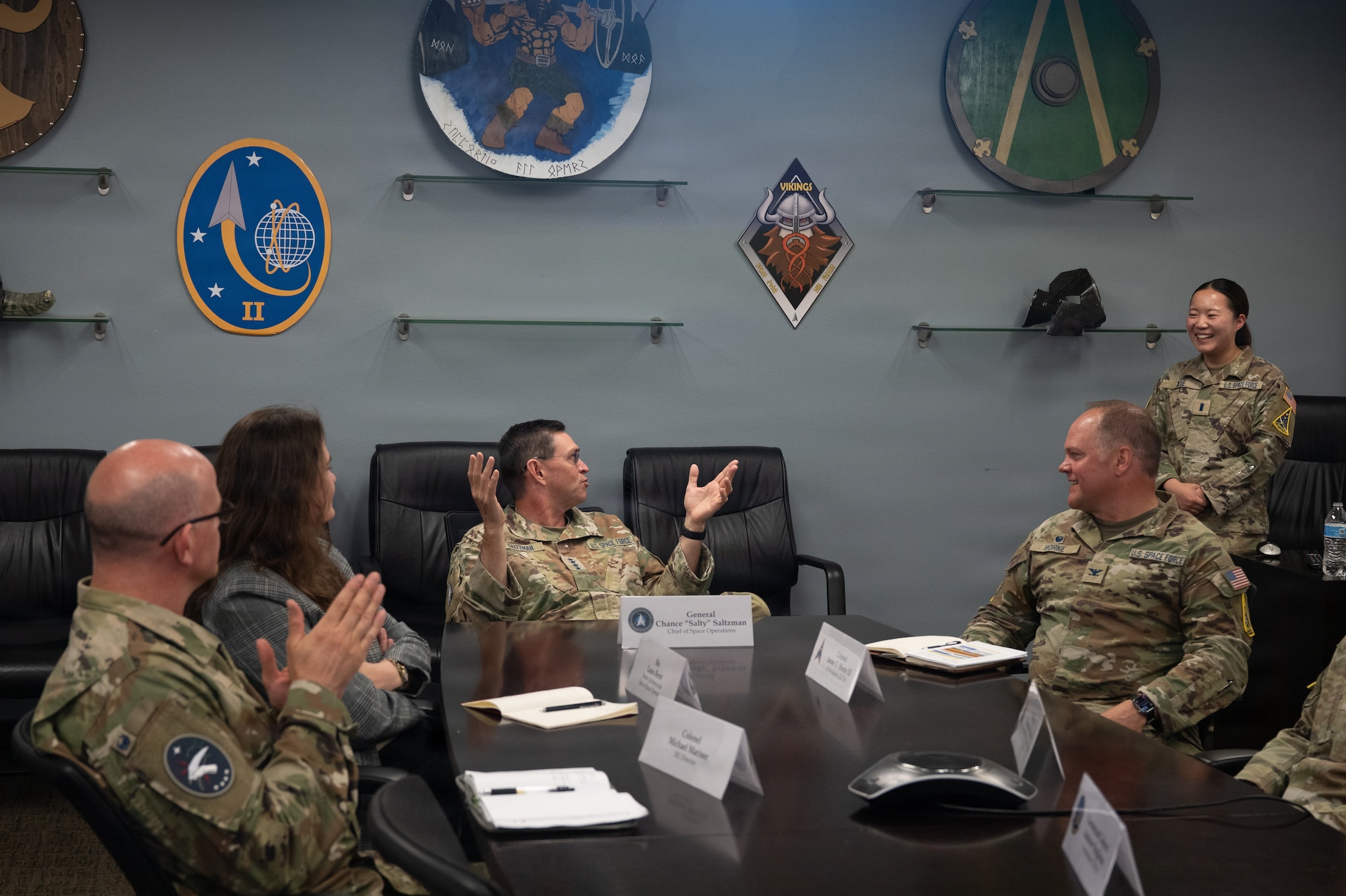Viking shield line the upper areas of a wall, a group of military members and civilians sit around a conference table as a general with hands raised palms up and away from his face in a shrug cracks a joke causing everyone to smile.
