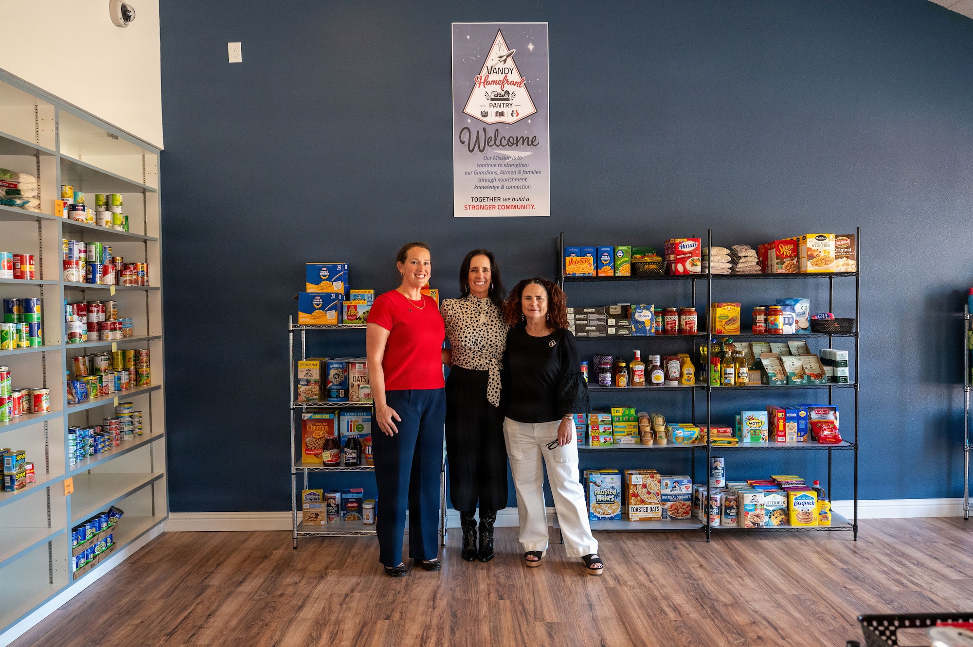 In a food pantry three women pose together by a wire rack filled with food.