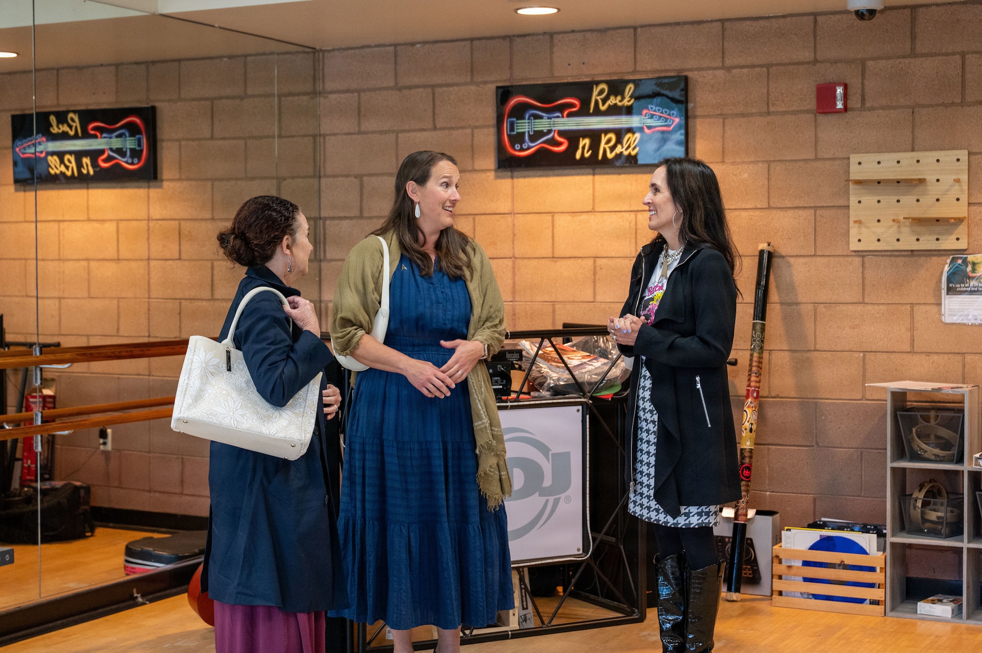 Three women talk in a music studio with a neon sign shaped like a guitar hanging in the background above their heads.