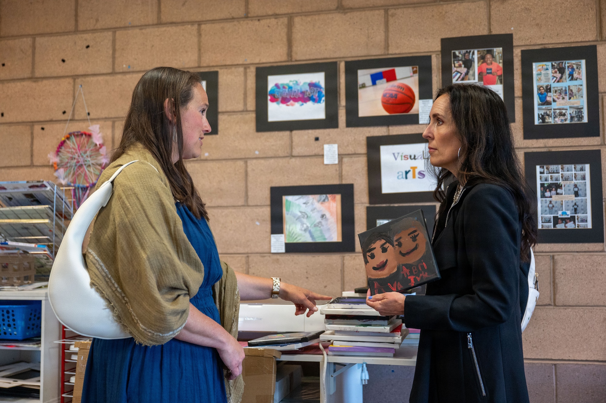 Two people go through paintings children made in a studio with painting on the walls.