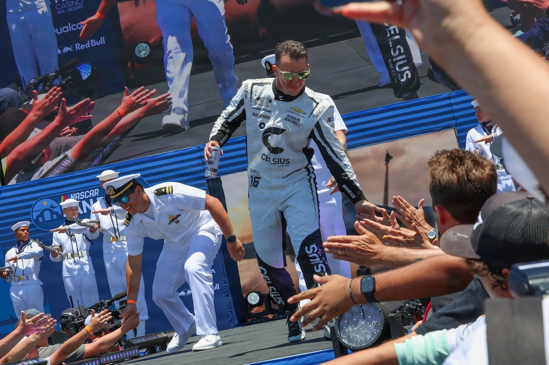 A man wearing a motorsports driving suit walks on a stage and slaps hands with members of a cheering crowd as sailors in dress uniforms walk and stand in formation behind him.
