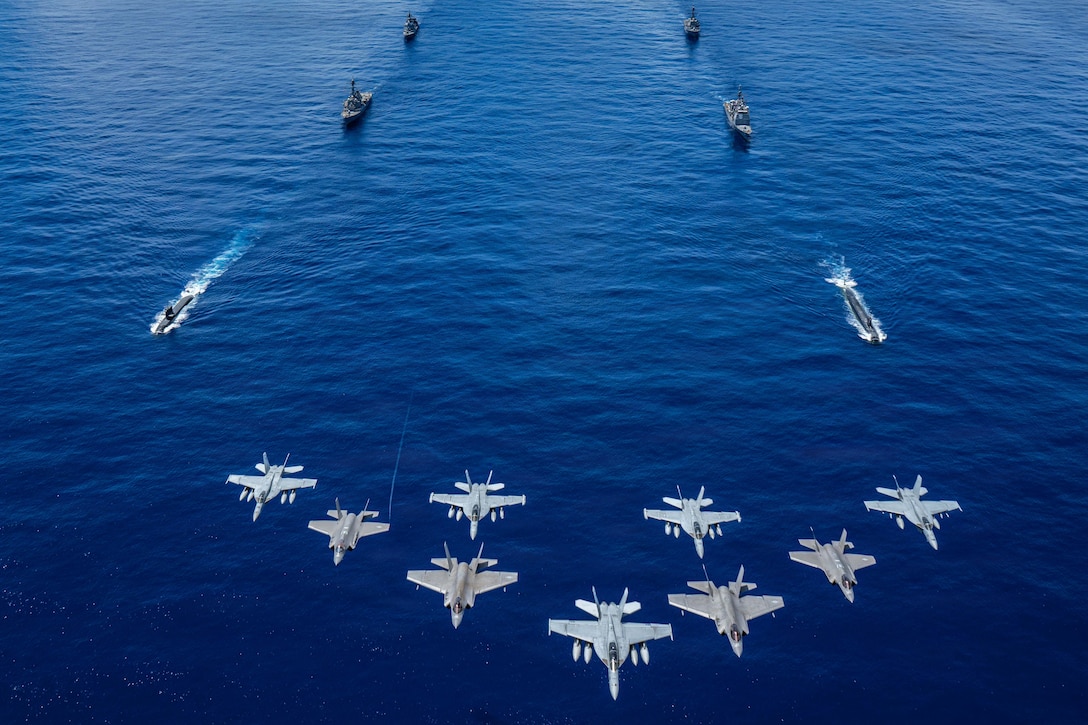 Nine aircraft fly over four ships and two submarines sailing in formation at sea.