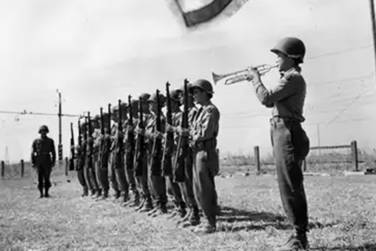 A black and white photo shows men in military uniforms standing in formation with rifles at their sides as another man in similar attire plays a bugle.