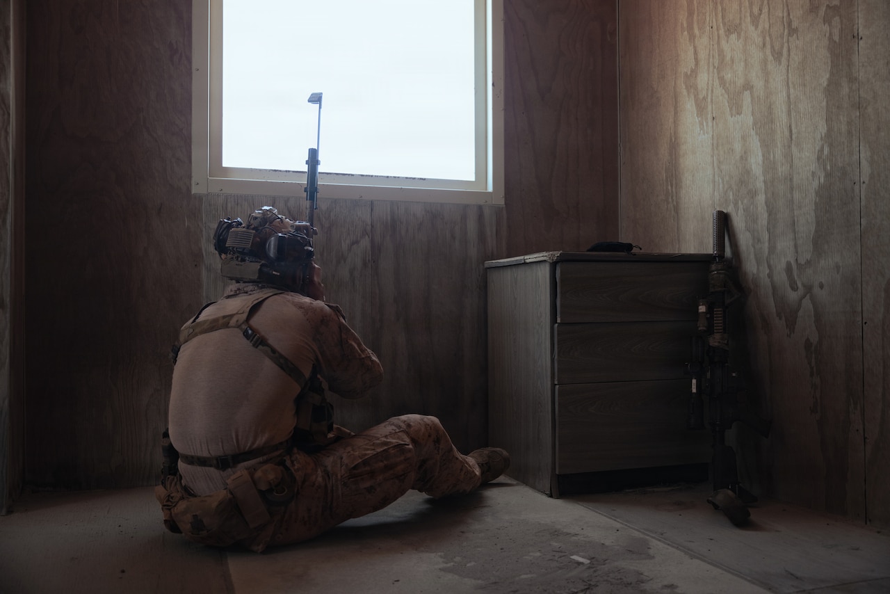 A person in a camouflage military uniform sits on the floor just below a window in a dimly lit room with a rifle in hand.