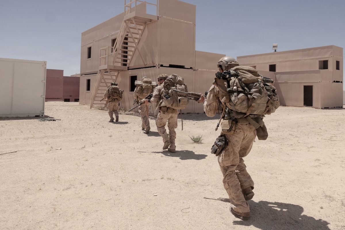 Four people in camouflage military uniforms walk across a desert terrain toward rows of empty shipping container buildings while carrying rifles.