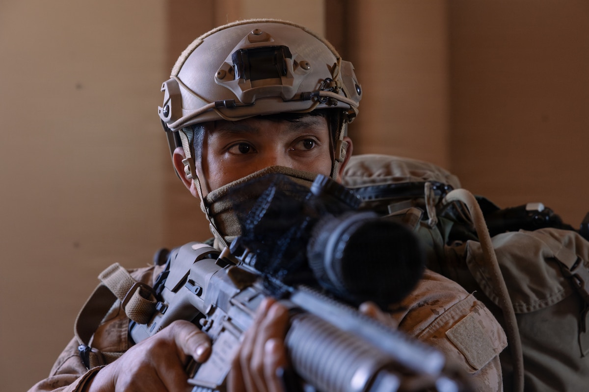 A man wearing a camouflage military uniform and a face mask looks to his left while holding a military rifle.