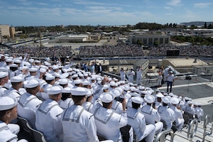 Secretary of War Pete Hegseth addresses sailors in white uniforms sitting in bleachers at a racetrack.