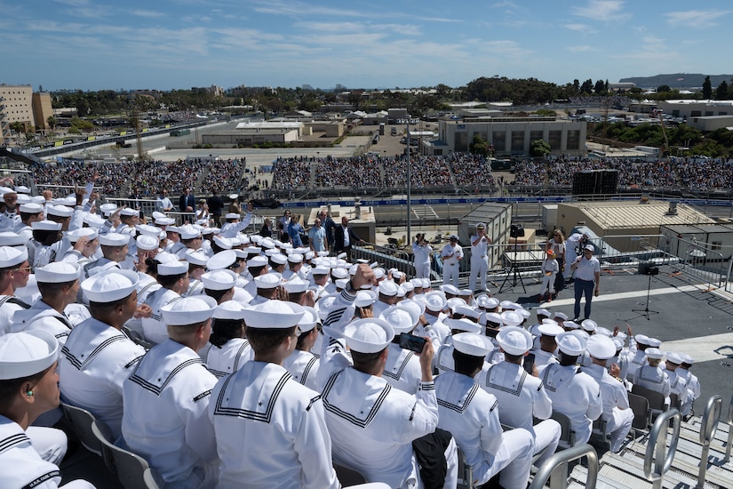 Secretary of War Pete Hegseth addresses sailors in white uniforms sitting in bleachers at a racetrack.