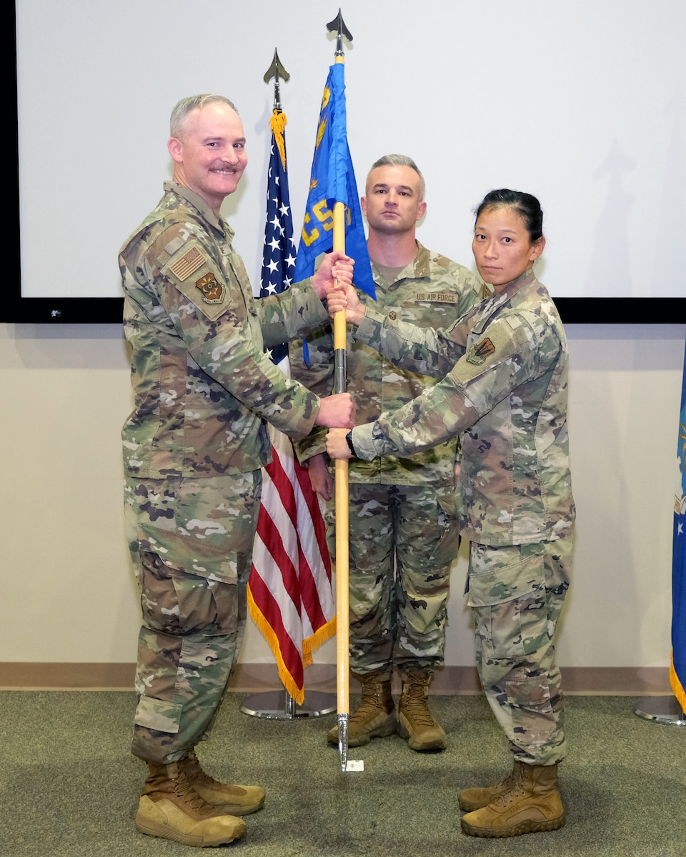 photo of three USAF Airmen standing on a stage, two Airmen are holding a unit flag in between them