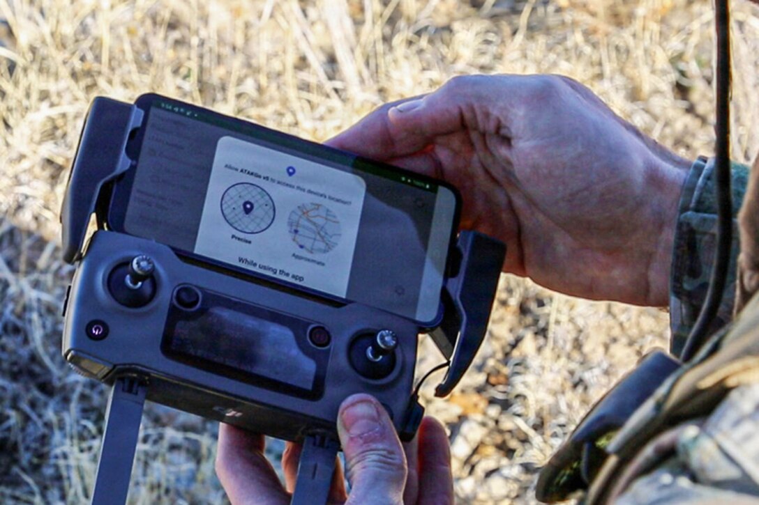 A soldier's hands hold a communication device with info on a screen with parched grass in the background.