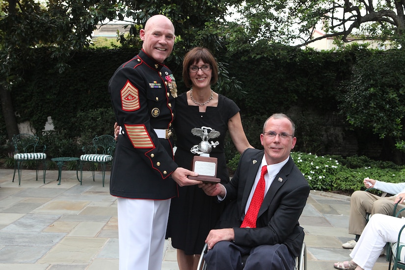 From left, Sgt. Maj. of the Marine Corps and Evening Parade host, Micheal P. Barrett; Rosemarie Cooper; and the Parade guest of honor, Dr. Rory Cooper, pose for a photo following a gift exchange during the Parade reception at Marine Barracks Washington in Washington, D.C.