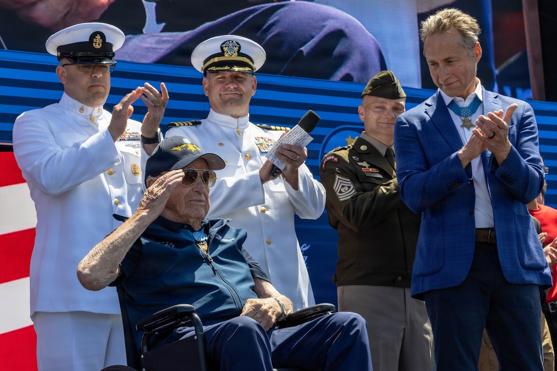 A veteran using a wheelchair salutes as a fellow veteran and other sailors in ceremonial dress clap in front of a patriotic banner.