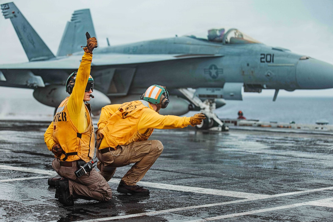 Two sailors wearing yellow vests and green helmets kneel on a wet flight deck while signaling to an aircraft.